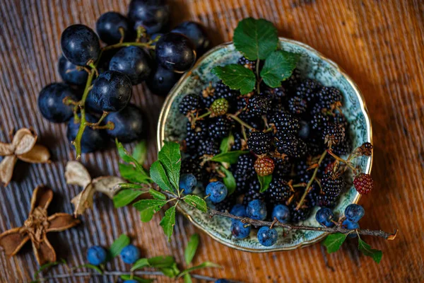 Üst manzara böğürtlenleri ahşap bir masada ve küçük bir tabakta mavi böğürtlen üzümleri. Yeşil yapraklar ve kuru çiçekler. Yüzeyin derinliği. Karanlık, bulanık bir arkaplan. Boşluğu kopyala.