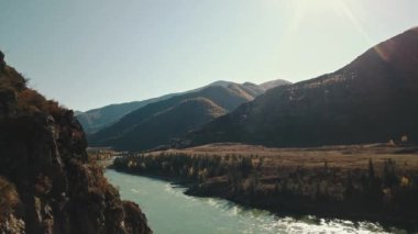 Sakin dağ nehri Katun 'un havadan görünüşü. Altai Bölgesi 'nin vahşi doğasının sonbahar manzarası: dağlar, ormanlar, tarlalar, kırsal bir yürüyüş yolu, güneş ışınları. Kamera uçuyor.