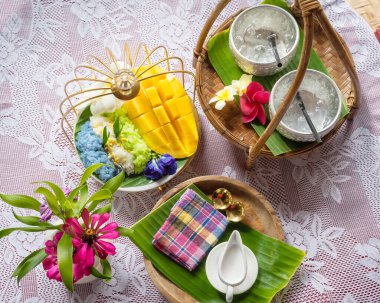 Muz yaprağında hindistan cevizi sütüyle karıştırılmış mangolu pirinç, ahşap masada beyaz seramik tabak ve beyaz giysi örtüsü Tayland 'da yaygın olarak kullanılan geleneksel bir tatlıdır. Meyve tatlısı..