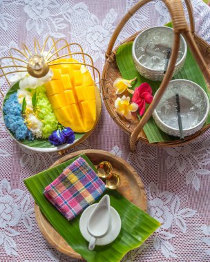 Muz yaprağında hindistan cevizi sütüyle karıştırılmış mangolu pirinç, ahşap masada beyaz seramik tabak ve beyaz giysi örtüsü Tayland 'da yaygın olarak kullanılan geleneksel bir tatlıdır. Meyve tatlısı..