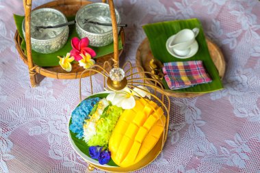 Muz yaprağında hindistan cevizi sütüyle karıştırılmış mangolu pirinç, ahşap masada beyaz seramik tabak ve beyaz giysi örtüsü Tayland 'da yaygın olarak kullanılan geleneksel bir tatlıdır. Meyve tatlısı..