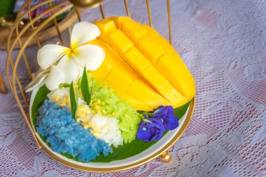 Muz yaprağında hindistan cevizi sütüyle karıştırılmış mangolu pirinç, ahşap masada beyaz seramik tabak ve beyaz giysi örtüsü Tayland 'da yaygın olarak kullanılan geleneksel bir tatlıdır. Meyve tatlısı..