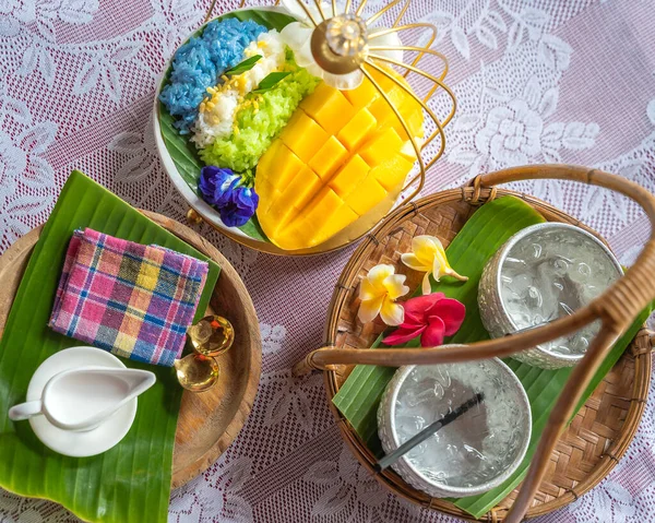 Muz yaprağında hindistan cevizi sütüyle karıştırılmış mangolu pirinç, ahşap masada beyaz seramik tabak ve beyaz giysi örtüsü Tayland 'da yaygın olarak kullanılan geleneksel bir tatlıdır. Meyve tatlısı..