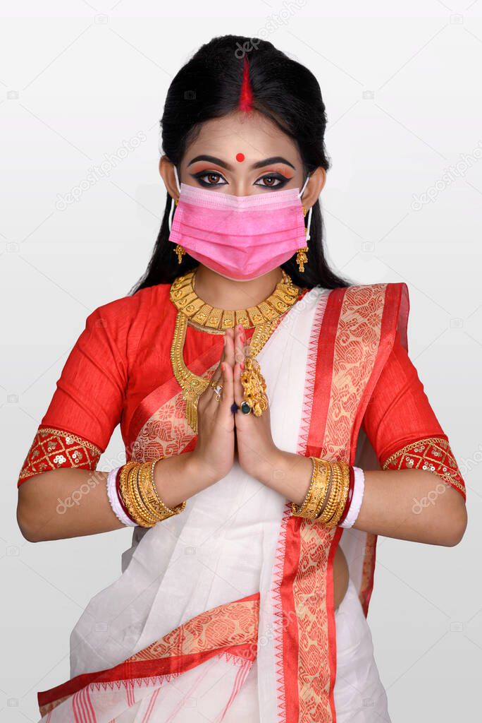 Portrait of pretty young Indian girl wearing traditional Indian saree, gold jewellery, bangles and mask standing in front of white background. Maa Durga agomoni shoot concept.
