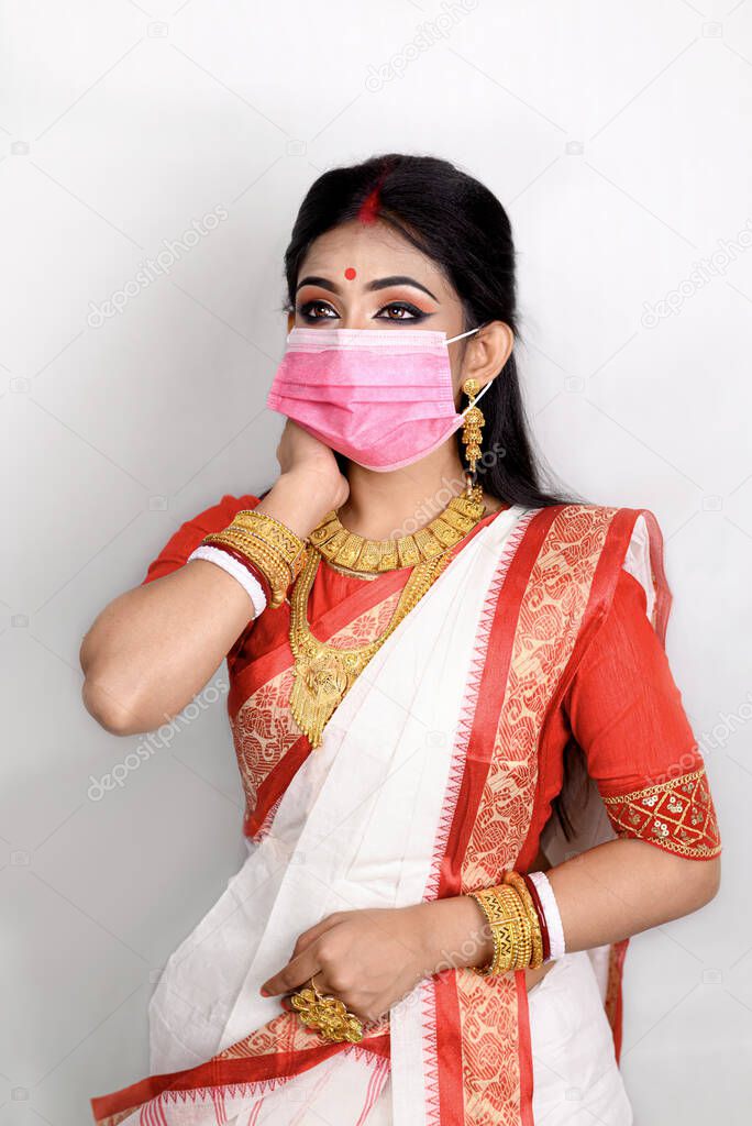 Portrait of pretty young Indian girl wearing traditional Indian saree, gold jewellery, bangles and mask standing in front of white background. Maa Durga agomoni shoot concept.