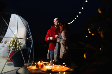 Ellerinde fincanlar olan sevgi dolu bir çift terasta glamping 'in önünde dururken birbirlerine sarılıyorlar. Parlayan çelenk masanın üzerinde yatıyor.