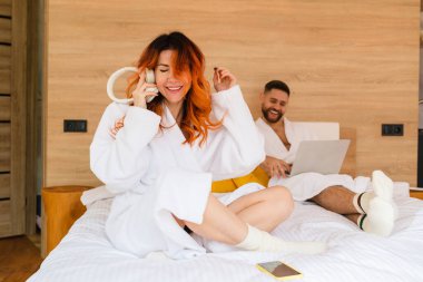 Bornozlu kızıl saçlı kadın kulaklıklı bir akıllı telefondan müzik dinliyor.