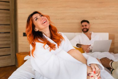 Beyaz bornozlu, kırmızı saçlı, gülümseyen genç bir kadın yatakta oturuyor..