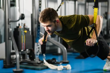 Spor giyimli güçlü bir adam trx fitness kayışlarıyla şınav çekiyor. Spor salonundaki genç fit adam, sağlıklı yaşam tarzı, modern çapraz ekipman ve spor egzersizleri için iç mekan..