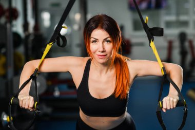 Ön manzara. Spor kıyafetleri içinde mükemmel vücutlu genç atletik bir kadın spor salonunda trx fitness kayışlarıyla şınav çekiyor.