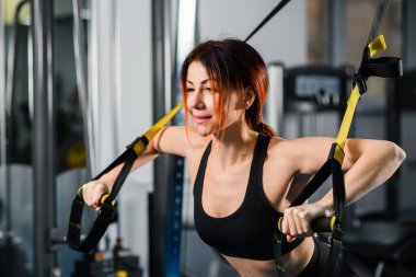 Spor kıyafetleri içinde mükemmel vücutlu genç atletik bir kadın spor salonunda trx fitness kayışlarıyla şınav çekiyor.