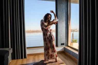 Odadaki büyük bir pencerenin önünde sabahlık giymiş çekici genç bir kadın dans ediyor. Güneşli bir sabah. Pencereden nehre bak.