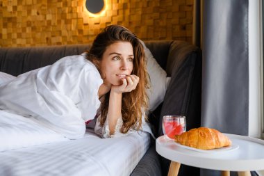 Çekici genç bir kadın sabahları yatakta kahvaltı yapıyor. Pencereden dışarı bakan kadın.