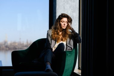 Rahatlamış bir kadın modern evdeki koltukta oturur ve panoramik penceredeki kapalı gözlerle güneşin tadını çıkarır.