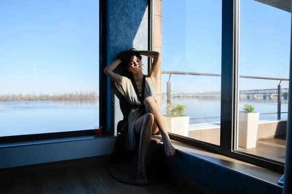 Sexy woman sit near panoramic window at luxury modern home interior with water river view with cup of tea, morning leisure