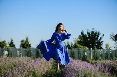 Lavanta tarlasında ilerleyen mavi elbiseli çekici genç bir kadın. Saçlar ve elbiseler rüzgarda savruluyor.