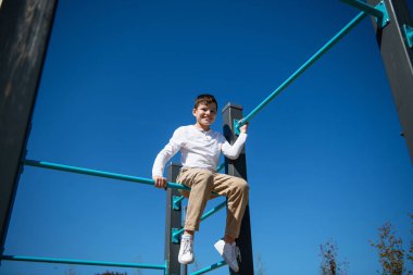 Genç bir çocuk yatay bir çubukta oturuyor. Alt manzara. Arka planda gökyüzü.