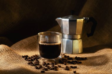 A transparent glass cup of coffee and a geyser coffee maker on a dark burlap background with coffee beans. Horizontal photo.
