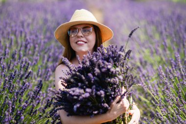 Hasır şapkalı genç bir kadın, lavanta tarlasında bir demet lavanta çiçeğiyle....