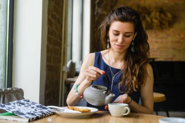 Genç bir kadın kahvecide otururken çaydanlıktan çay dolduruyor..