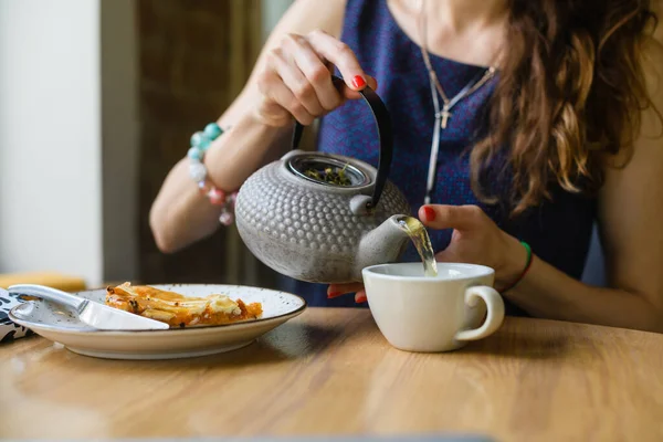 A young woman pours tea from a teapot while sitting in a coffee shop. Close-up.