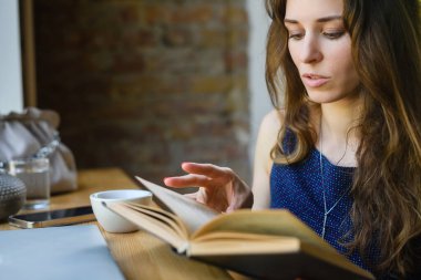 Attractive young girl reading a book while having breakfast in a coffee shop. Close-up.