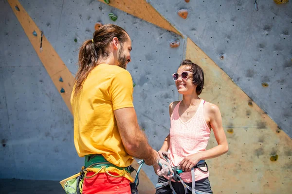 A man helps a young woman to put on her climbing equipment against the backdrop of a climbing wall outdoors.