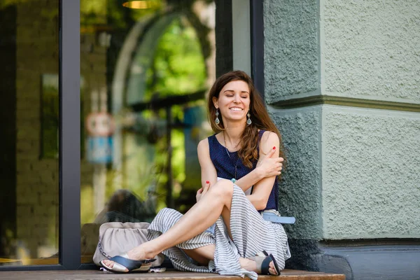 Young attractive woman sits cross-legged by a huge window outdoors. Young attractive woman sits cross-legged by a huge window outdoors. The girl hugs herself with her hands.