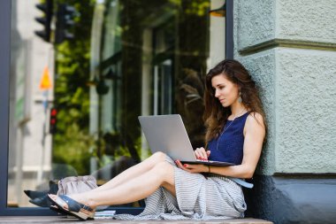 Young attractive woman with laptop sits on the windowsill outdoors. Huge window, showcase in the background.