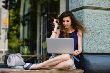 Young attractive woman with laptop sits on the windowsill outdoors. The girl straightens her hair with her hand.