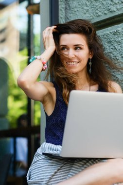 Young attractive woman with laptop sits on the windowsill outdoors. The girl straightens her hair with her hand.