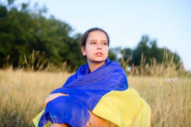 A teenage girl sits in tall wild grass on the slope of a ravine with a Ukrainian flag on her shoulders.