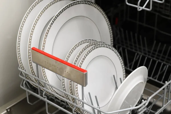 Open dishwasher with clean dishes in a white kitchen. Clean plates, cups and dishes. Nobody. Selective focus.