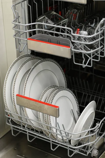 Open dishwasher with clean dishes in a white kitchen. Clean plates, cups and dishes. Nobody. Selective focus.