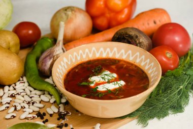 Traditional Ukrainian borscht with sour cream in a clay bowl. Ingredients for cooking borscht on a wooden cutting board.