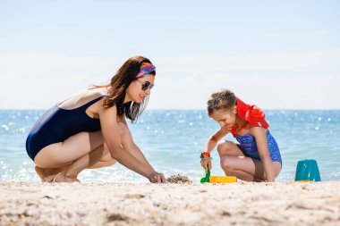 Anne ve okul çağındaki kızı mutlu aile tatili sırasında kumsalda kumla oynuyorlar, aktif bir çocukluk.