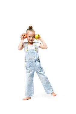 Kot pantolon giymiş şirin bir kız beyaz arka planda elmalarla oynaşıyor. Kulakları için elma tutan bir kız