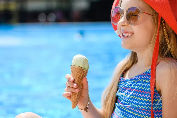 Happy little girl child sit with ice cream near hotel resort swimming pool in swimsuit and summer hat, family vacation