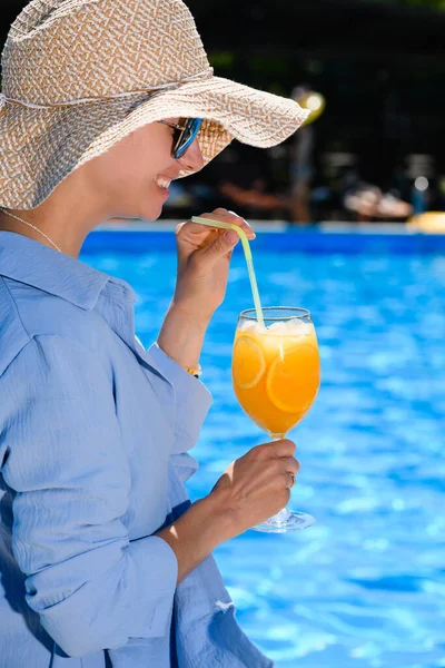 Happy smiling woman in sun glasses and hat drink citrus fresh cocktail near swimming pool, summer vacation vibes