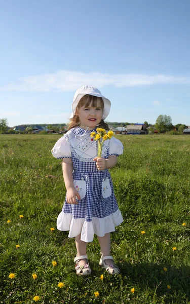 Happy little girl wearing dress holds yellow dandelions on green — Stock Photo, Image