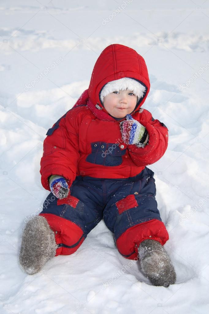 Little girl wearing red jumpsuit sits on snow and eats snow at w