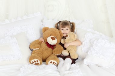 küçük kız Çift Kişilik Yatak ile çok sayıda yastıklar ve oyuncak ayılar üzerinde oturur.