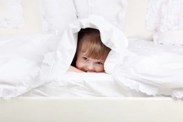 Little beautiful girl looks out from under warm blanket on bed. — Stock Photo, Image