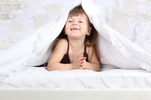 Little girl looks out from under warm blanket on double bed and — Stock Photo, Image