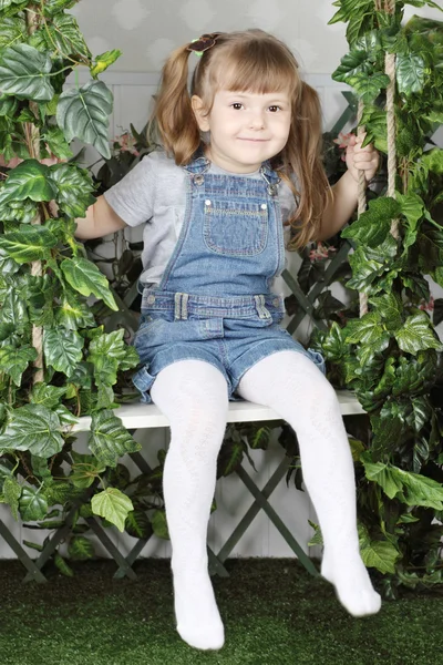 Happy little girl in denim jumpsuit sits on swing under green iv — Stock Photo, Image