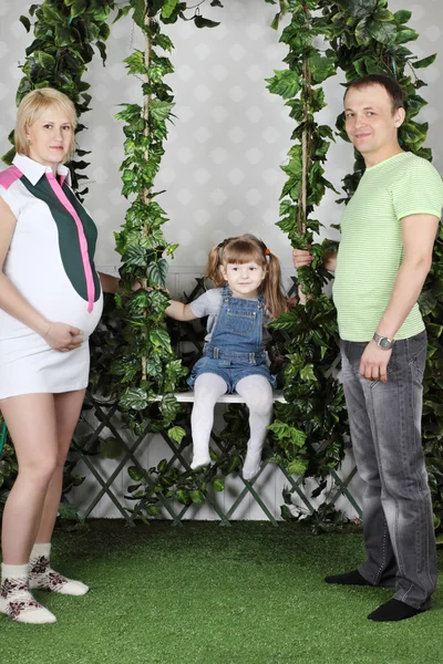 Malá holka sedí na houpačce pod zeleným břečťanem a otec a pregnan — Stock fotografie