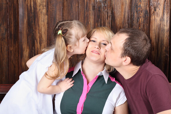 Отец и маленькая дочь целуют мать на скамейке рядом с деревянной
