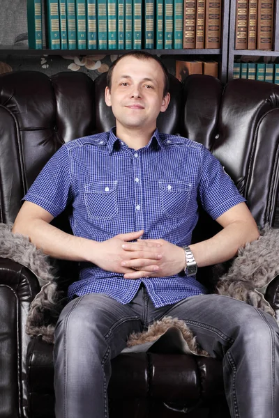 Young man sits in leather armchair next to shelves with lots of — Stock Photo, Image