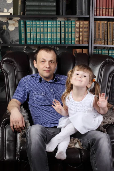 Man and her daughter sit in leather armchair next to shelves wit — Stock Photo, Image