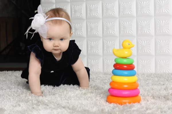 Weinig schattige baby in jurk kruipt op grijs zachte tapijt onder speelgoed. — Stockfoto